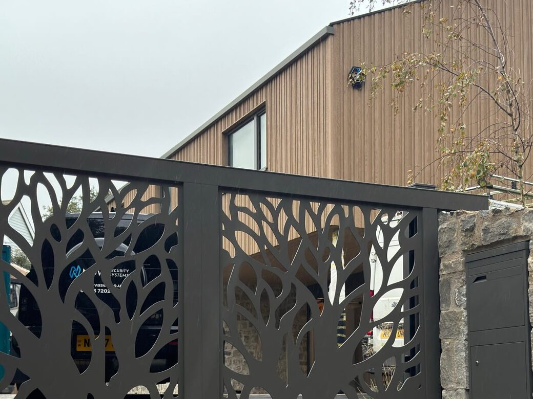 Custom waterjet-cut grey sliding gates with a tree pattern in front of a new-build timber-clad house in Kent