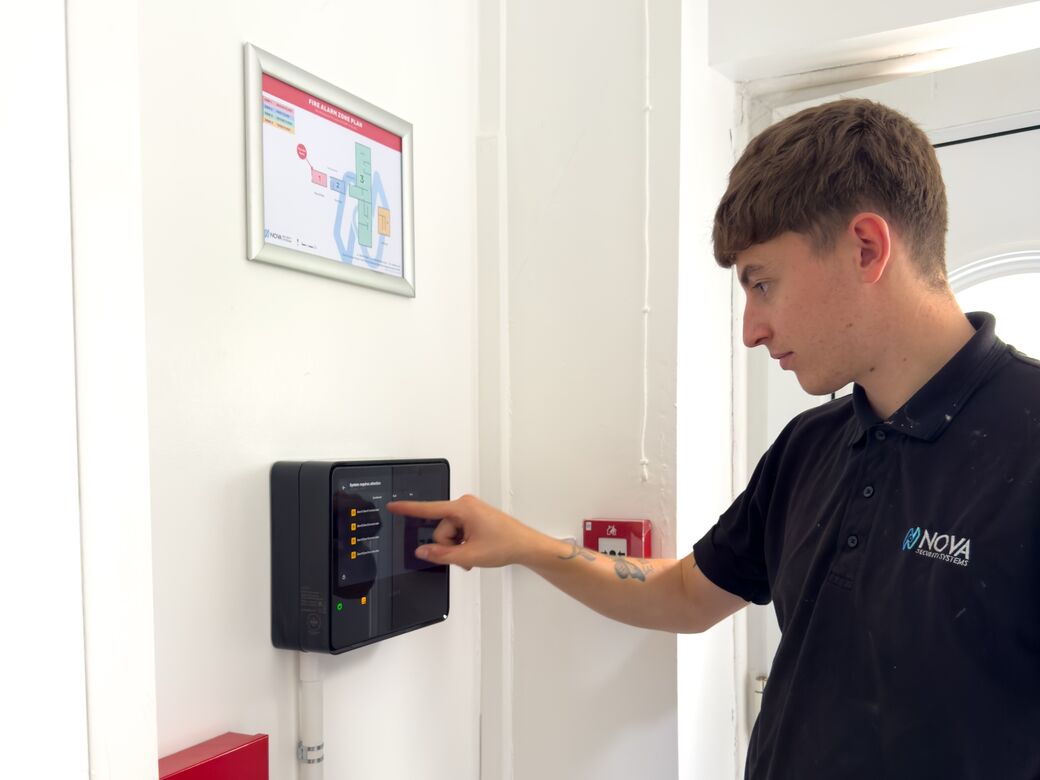 A Nova engineer commissioning an Ajax EN54 Fire Hub in a Woolwich HMO, with the framed zone plan above